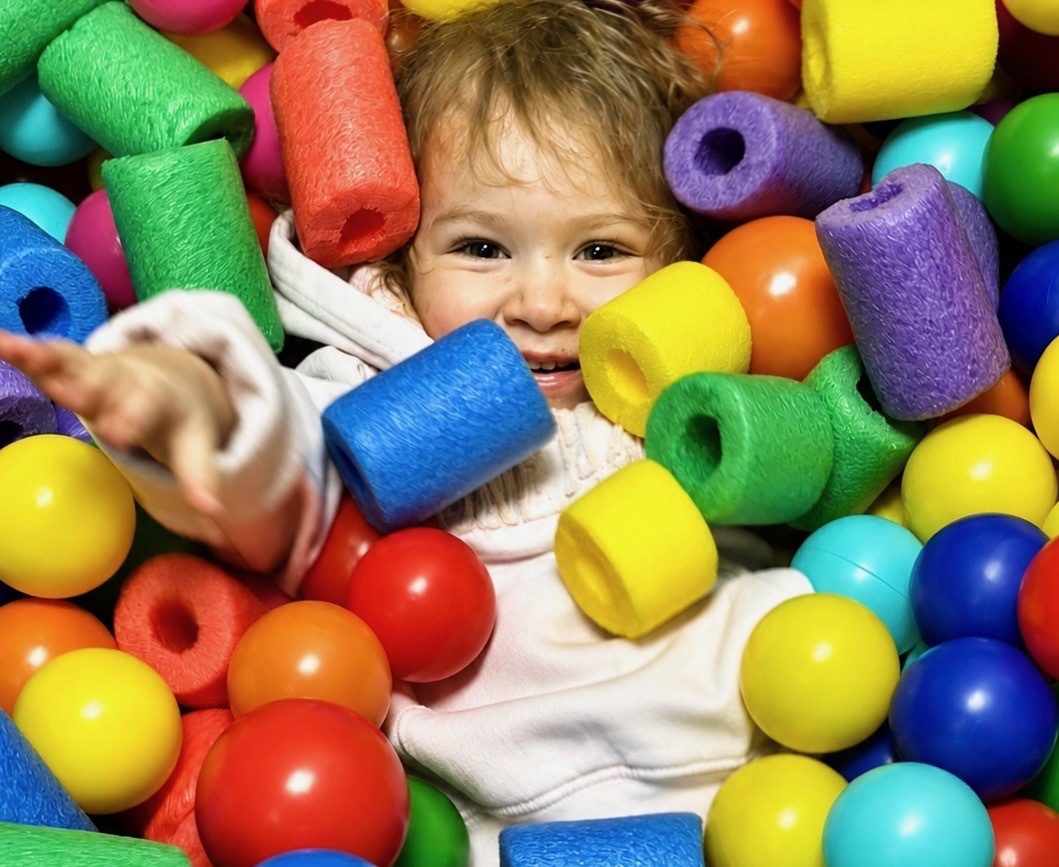 A toddler smiling and pointing while buried in colorful foam pool-noodles and balls.